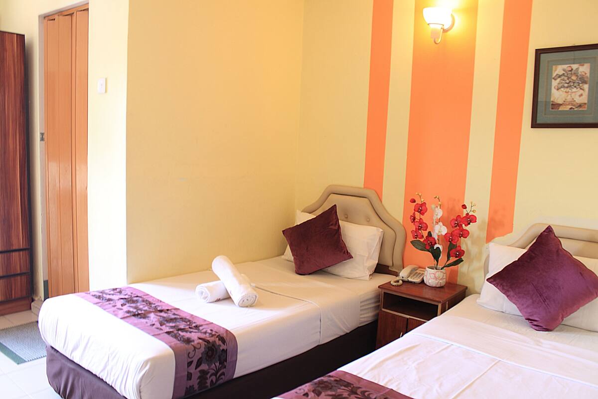 A hotel room with two single beds, a wooden wardrobe, a nightstand with a phone, and a wall-mounted light fixture.
