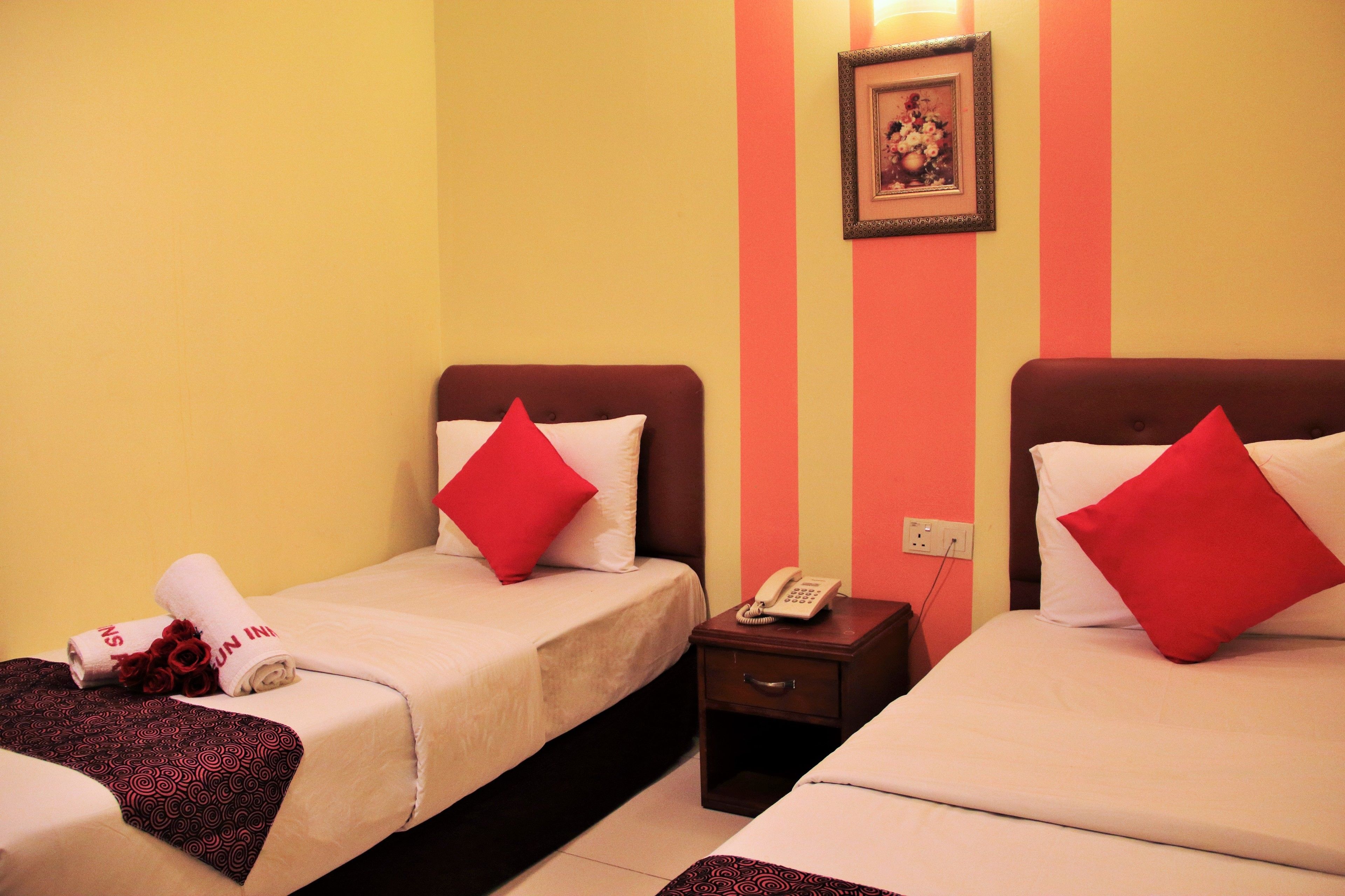 A hotel room with two beds, each with a red pillow, a bedside table with a phone, and a wall-mounted picture.