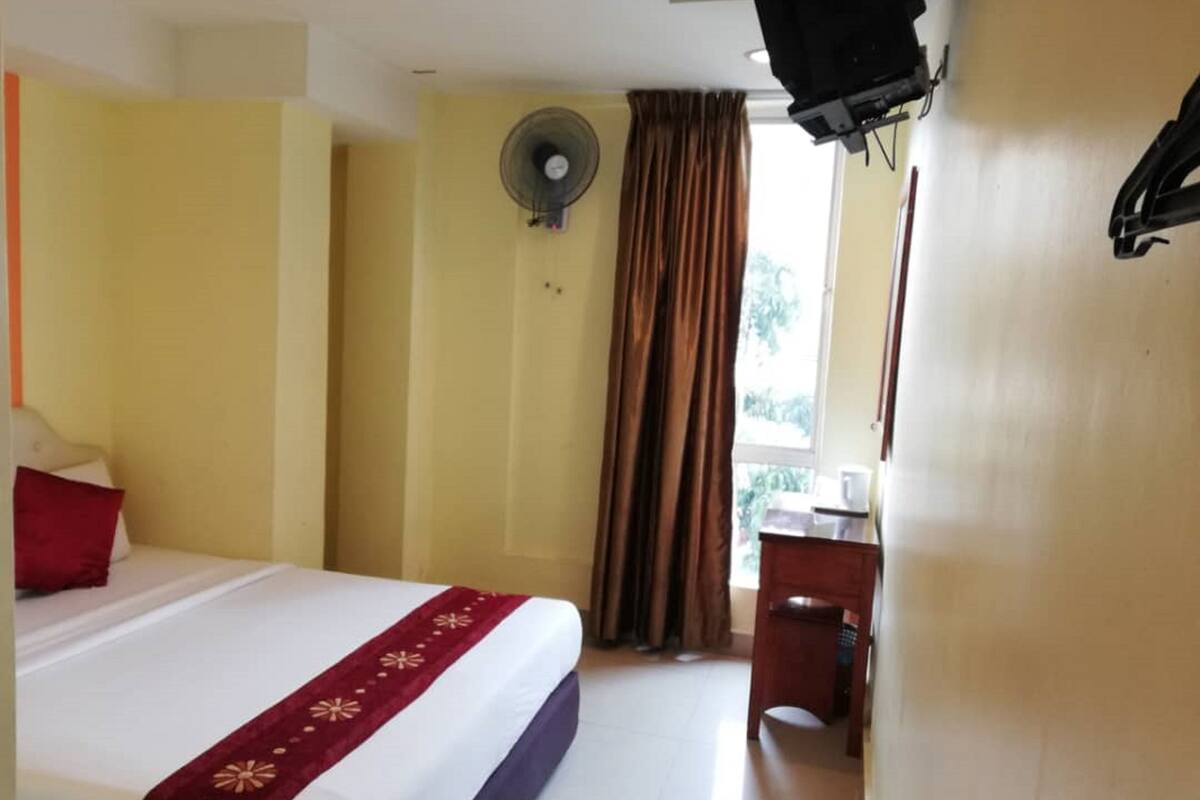 A hotel room with a bed, a desk, a fan, and a window with curtains.