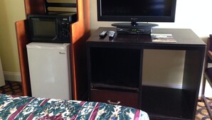 A compact room with a bed, a small refrigerator, a microwave, a television, and a remote control on a wooden cabinet.