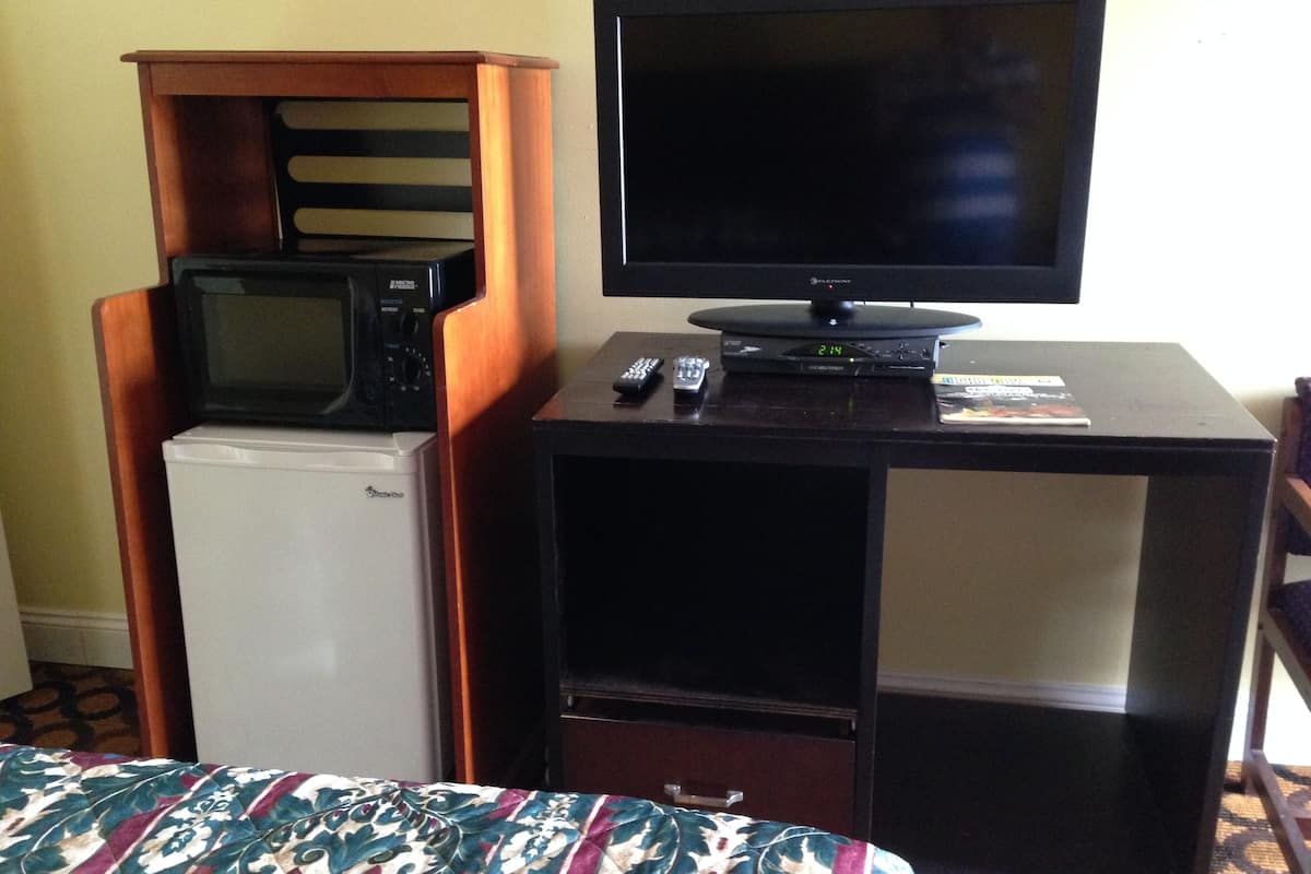 A compact room with a bed, a small refrigerator, a microwave, a television, and a remote control on a wooden cabinet.