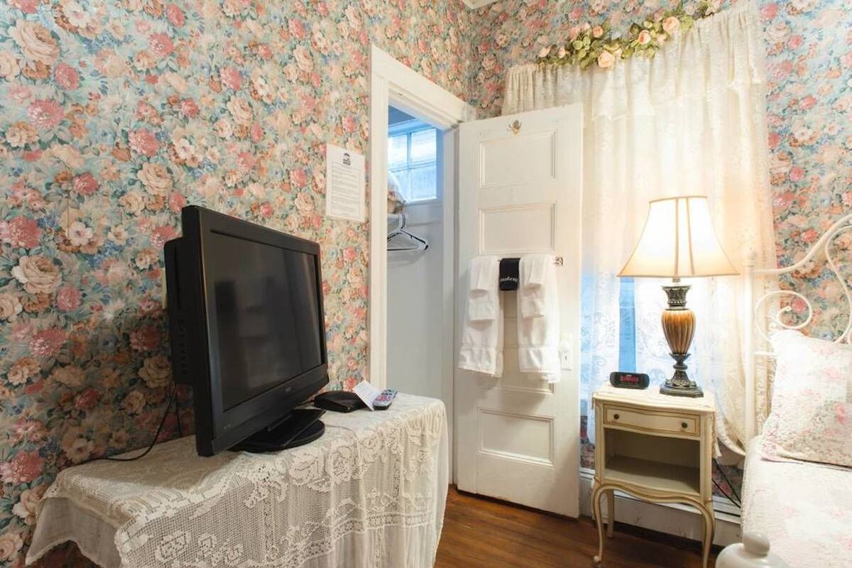 Una stanza con carta da parati a motivi floreali, una televisione su un tavolino, una lampada da comodino e una porta che dà su un'altra stanza.