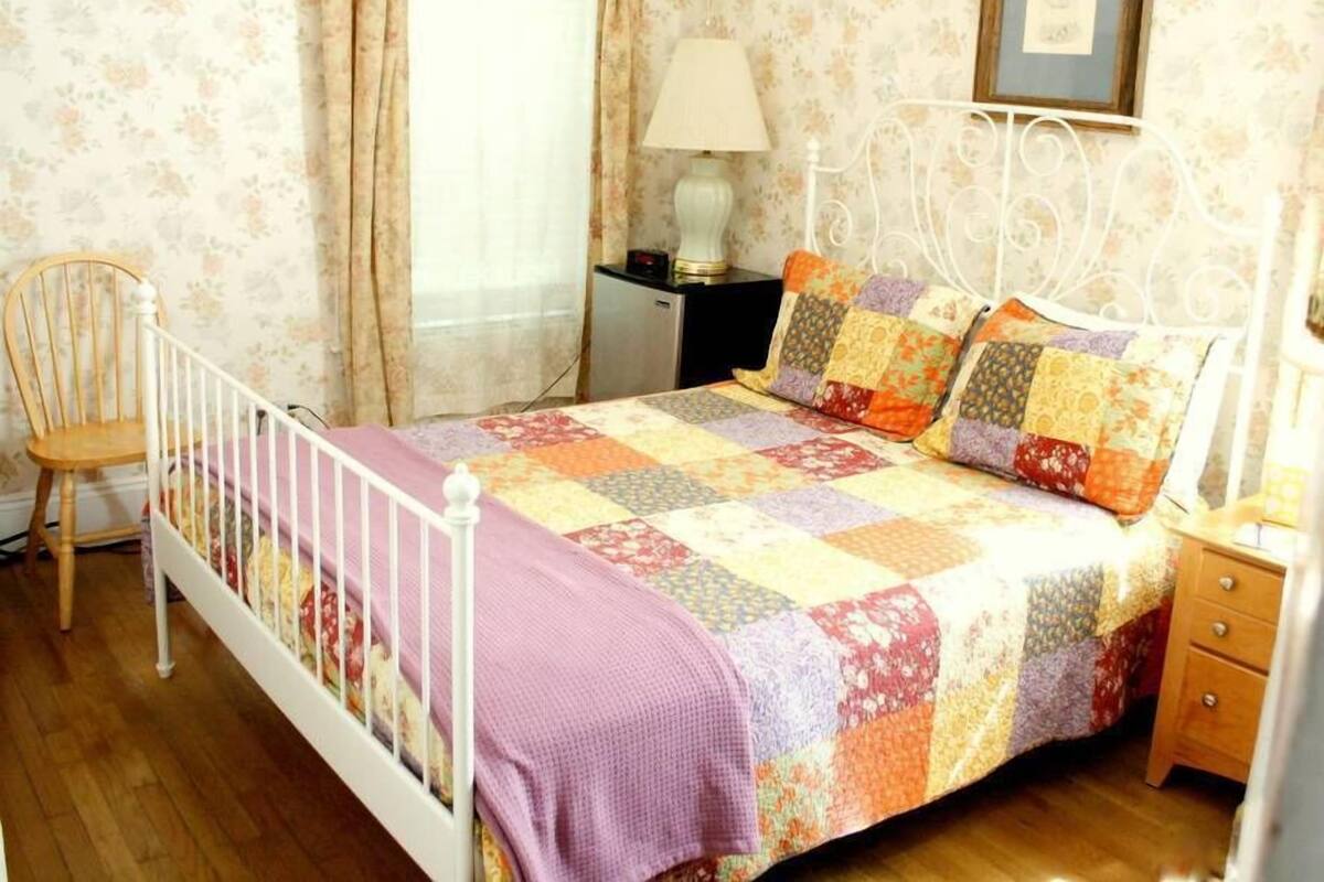 Una camera da letto con un letto con struttura in metallo bianco, una coperta colorata, una sedia di legno, un comodino, una lampada e un quadro appeso al muro.