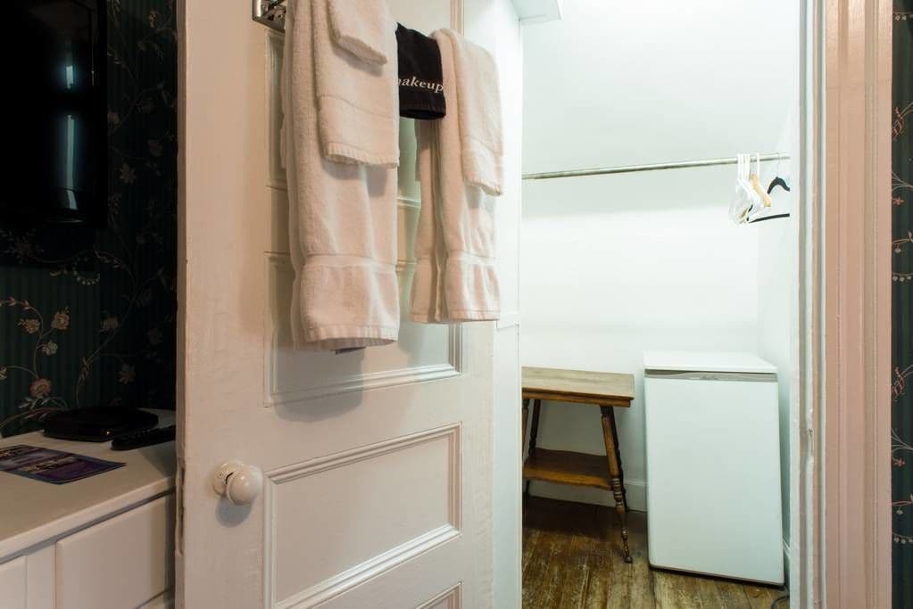 Ein kleiner, kompakter Raum mit einer weißen Tür, einem weißen Kühlschrank, einem Holzregal und einer schwarzen Tasche, die an der Tür hängt.