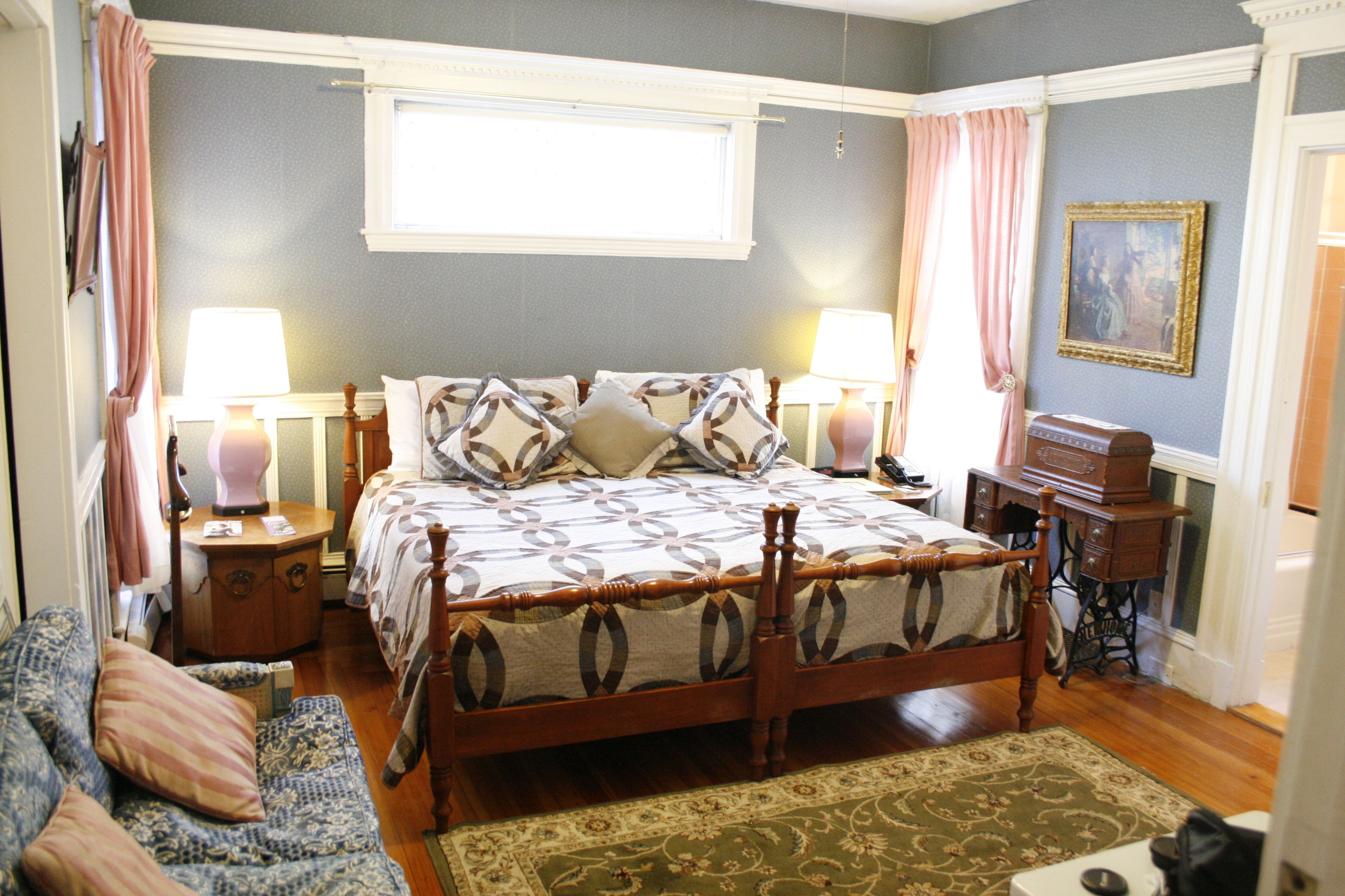 A bedroom with a wooden four-poster bed, two bedside tables, a nightstand, a dresser, a window with curtains, and a painting on the wall.