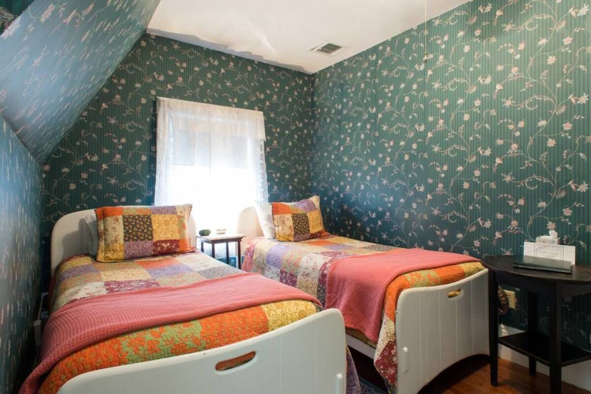 Una stanza con due letti, ognuno con una coperta colorata e un poggiatesta bianco. Si vede un tavolino con una lampada e una finestra con una tenda leggera.