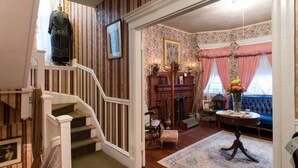 A grand staircase leading to a living room with a fireplace, a blue sofa, and a table with a vase of flowers. - Coolidge Corner Guest House (Brookline)