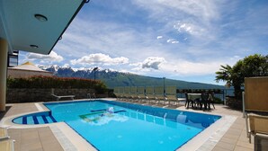 Piscina con sillas de descanso y mesas, rodeada por un muro de piedra y vistas a montañas nevadas.