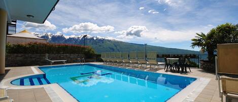 A swimming pool with lounge chairs and tables, surrounded by a stone wall and a view of snow-capped mountains.