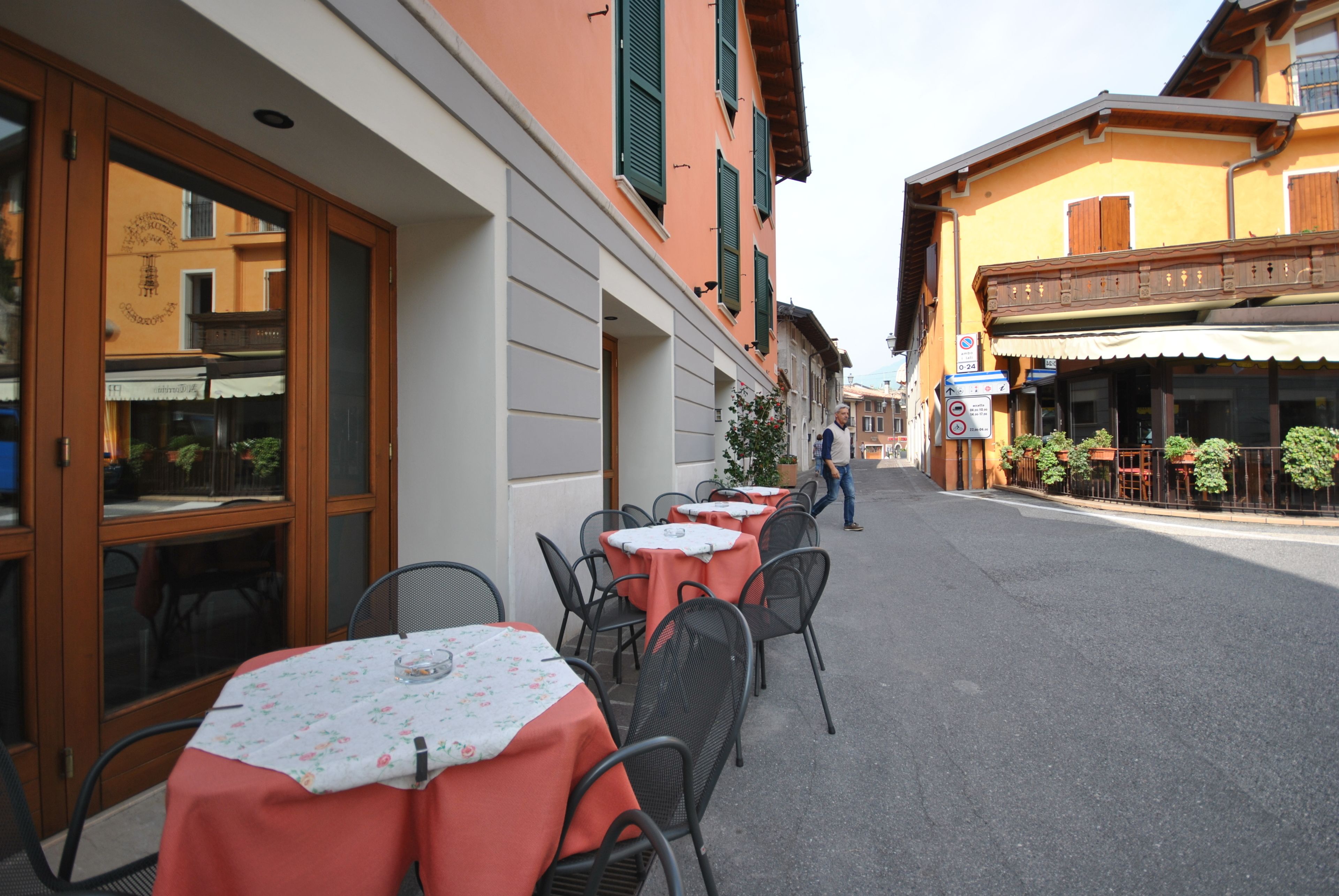 An outdoor dining area with tables and chairs, a street in front, and buildings with balconies and shutters.