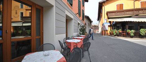An outdoor dining area with tables and chairs, a street in front, and buildings with balconies and shutters.