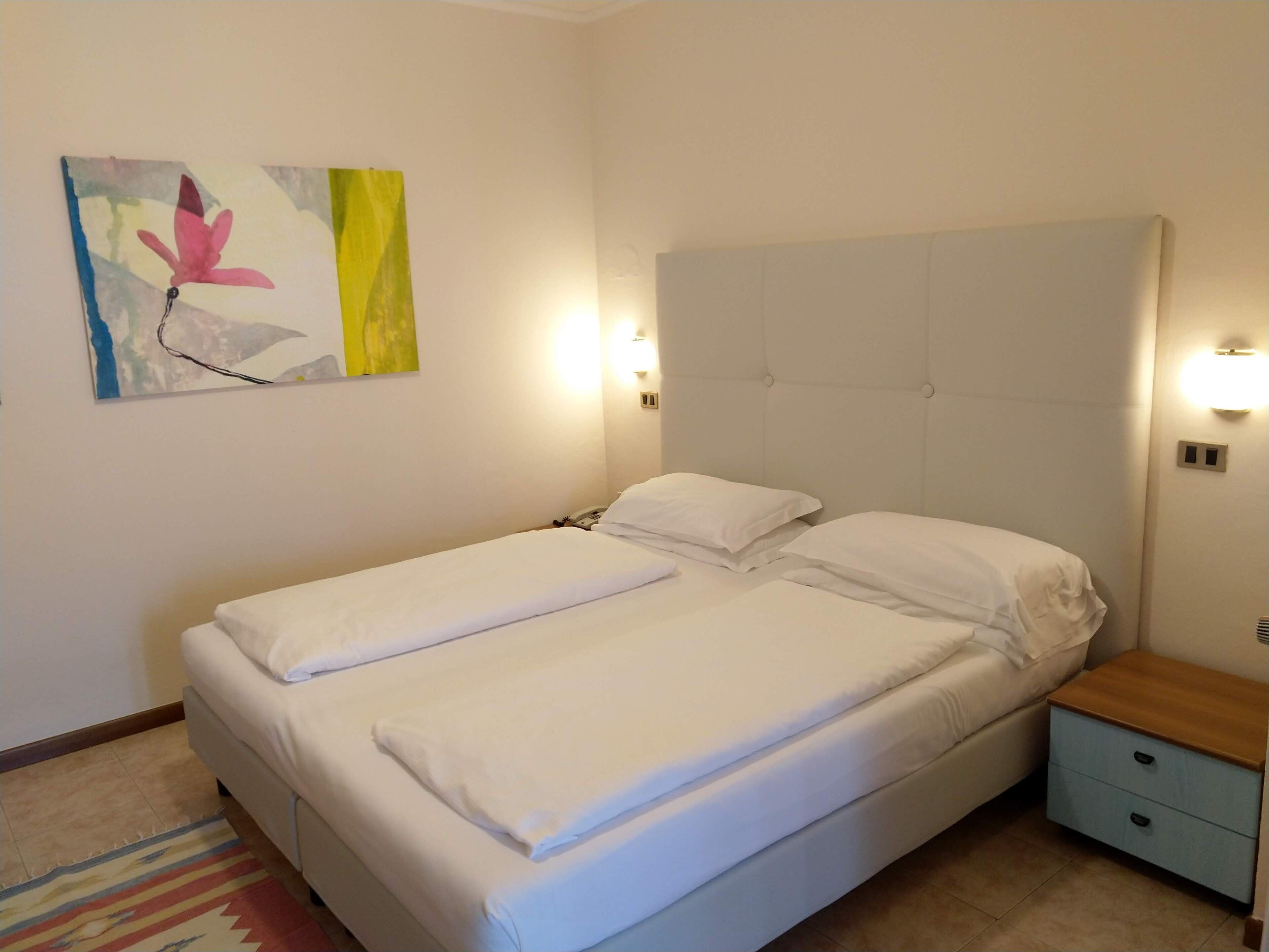 A double bed with white linens, a headboard, bedside lamps, a nightstand, and a colorful rug.