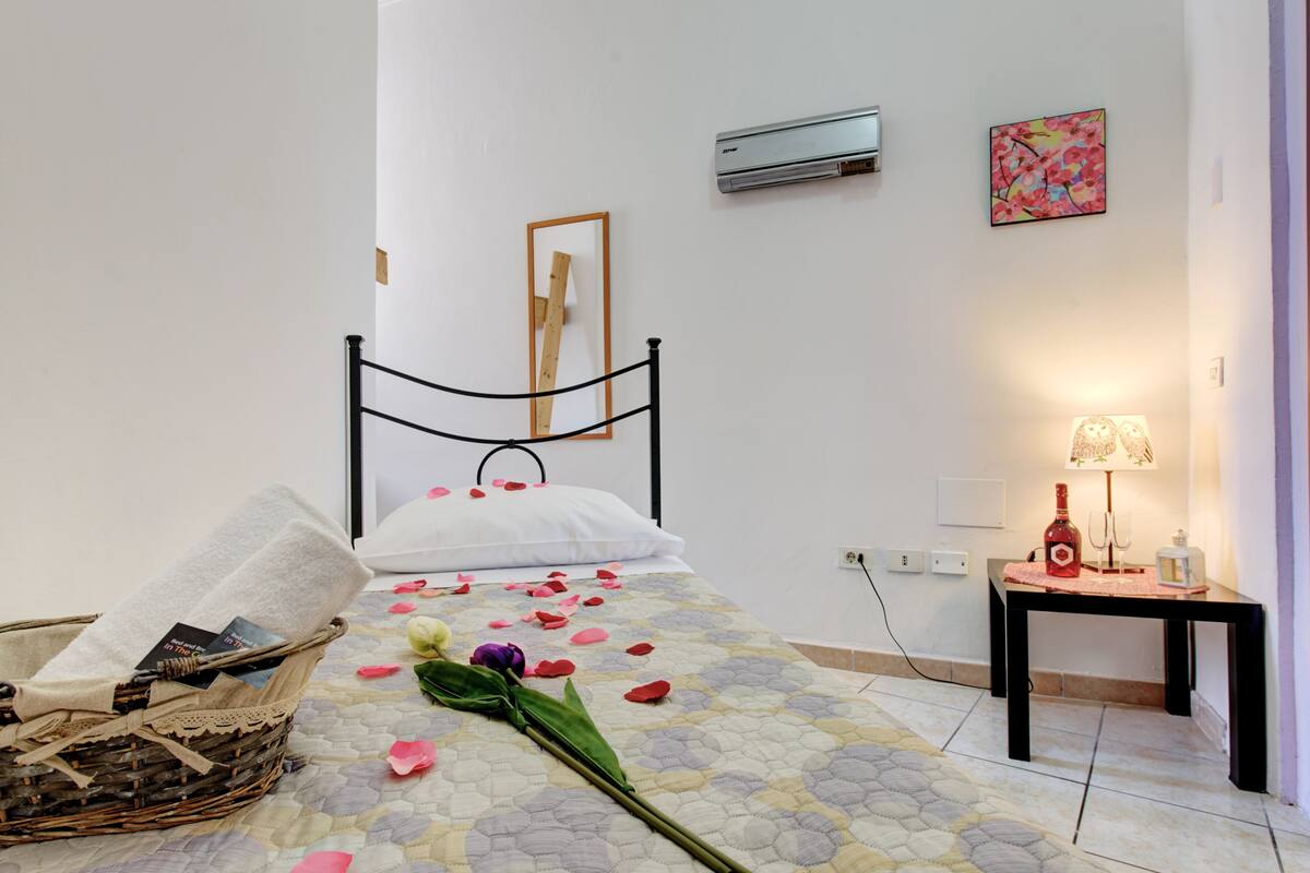 Une chambre à coucher bien rangée, avec un lit, une table de chevet dotée d’une lampe, une petite table sur laquelle se trouve une bouteille, et un panier en osier.