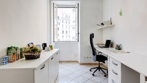 Ein kleiner, heller Raum mit einem weißen Schreibtisch, einem schwarzen Bürostuhl und einem Fenster mit Blick auf Gebäude.