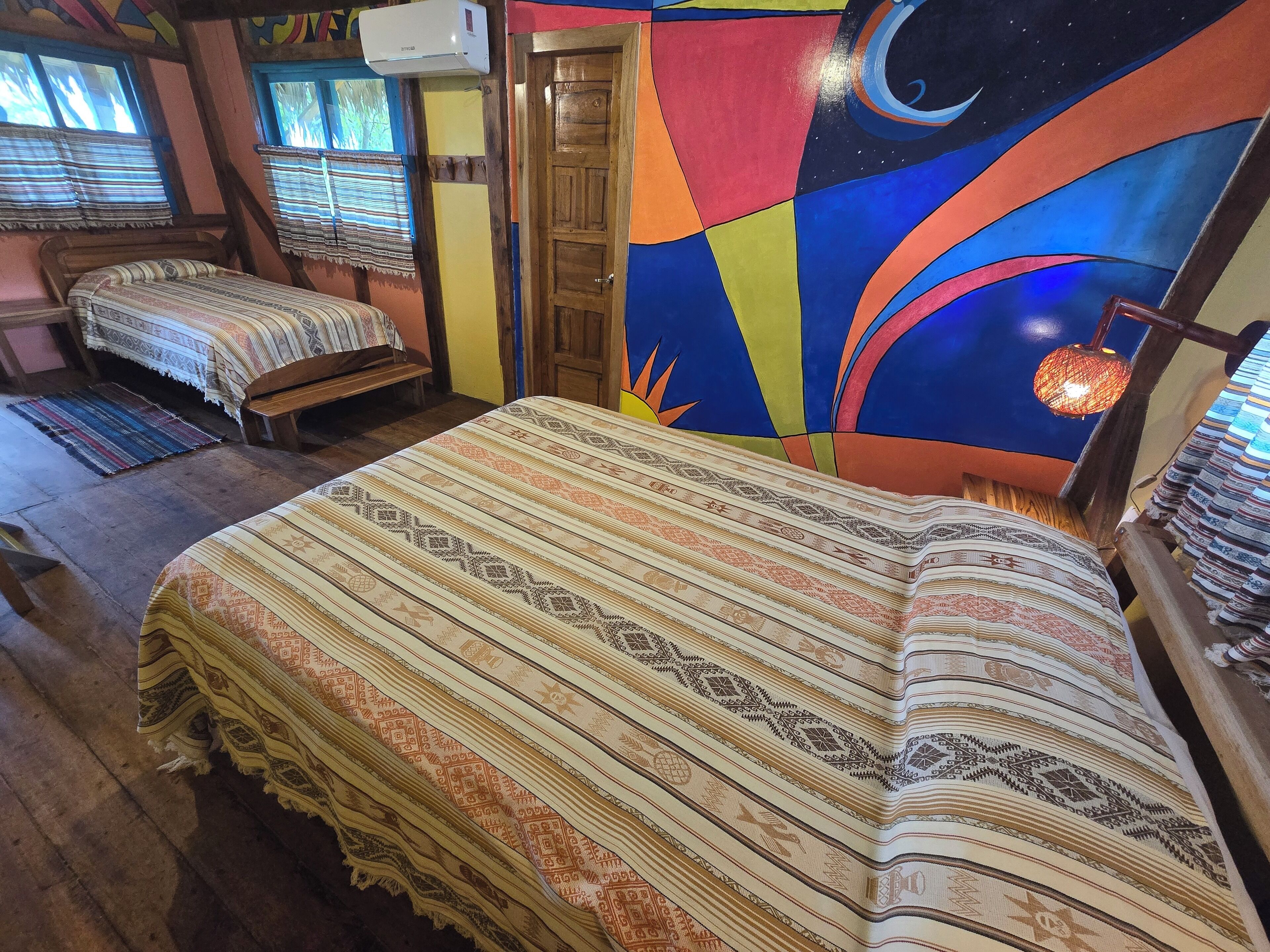 A room with a large bed, a wooden bench, a colorful wall mural, and a small table with a lamp.
