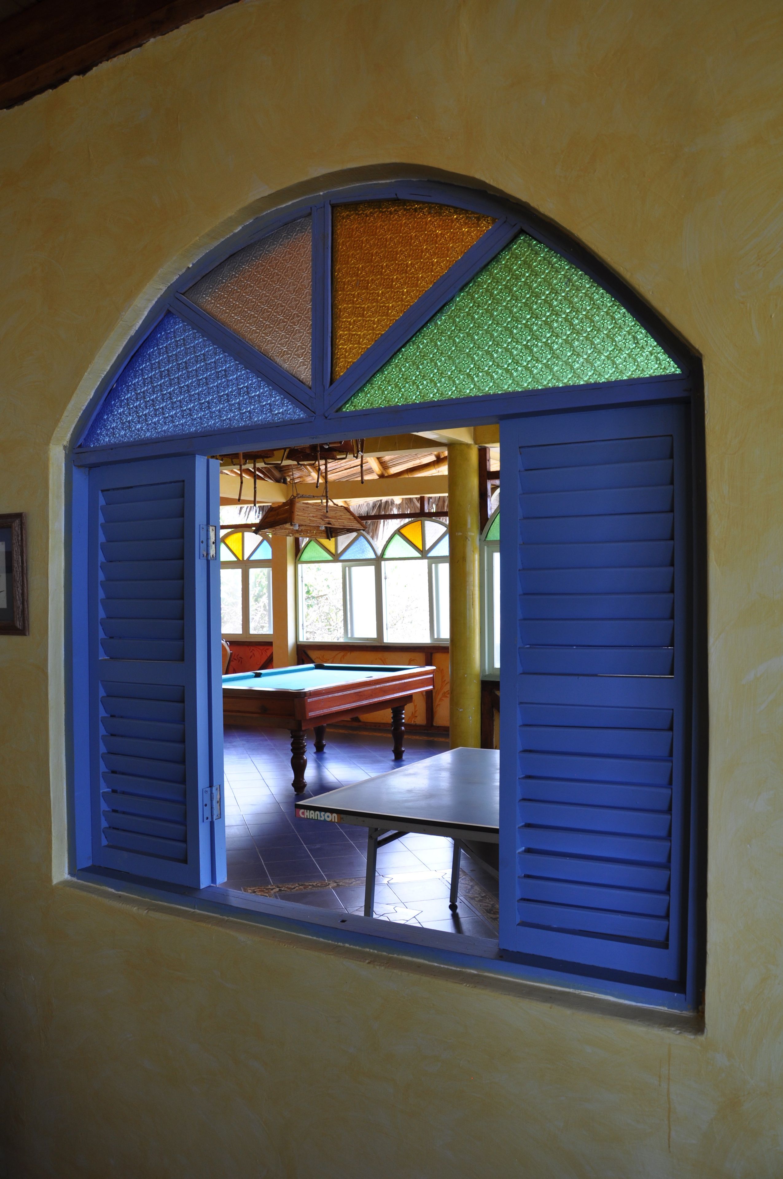 A room with a pool table, a window with blue shutters, and a view of an outdoor area with a thatched roof.