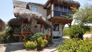 Un edificio de dos pisos con techo de paja, balcones de madera y un jardín con plantas tropicales.