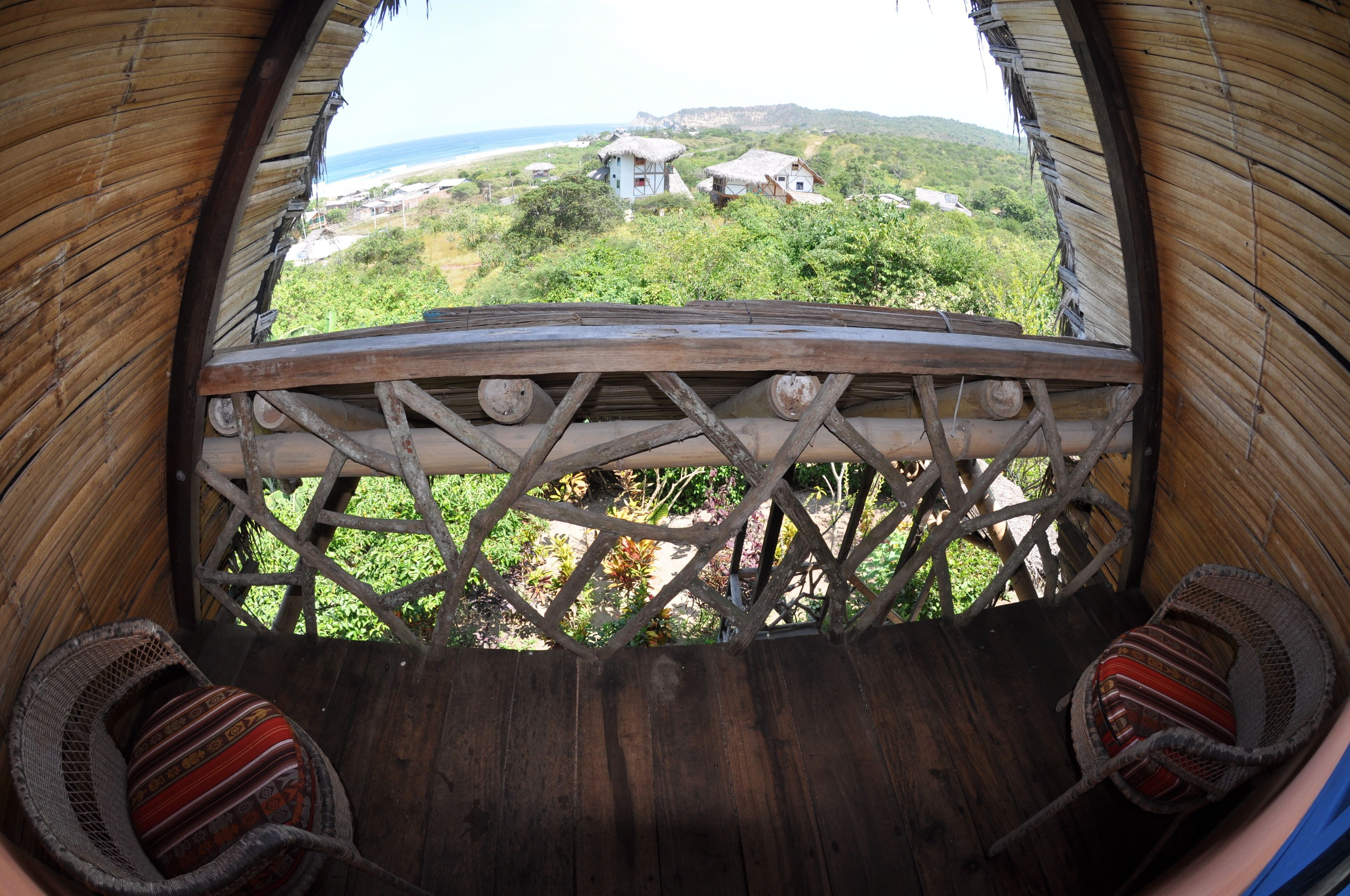Vista desde una construcción con techo de paja y balcón de madera, sobre una zona costera con edificios y vegetación.