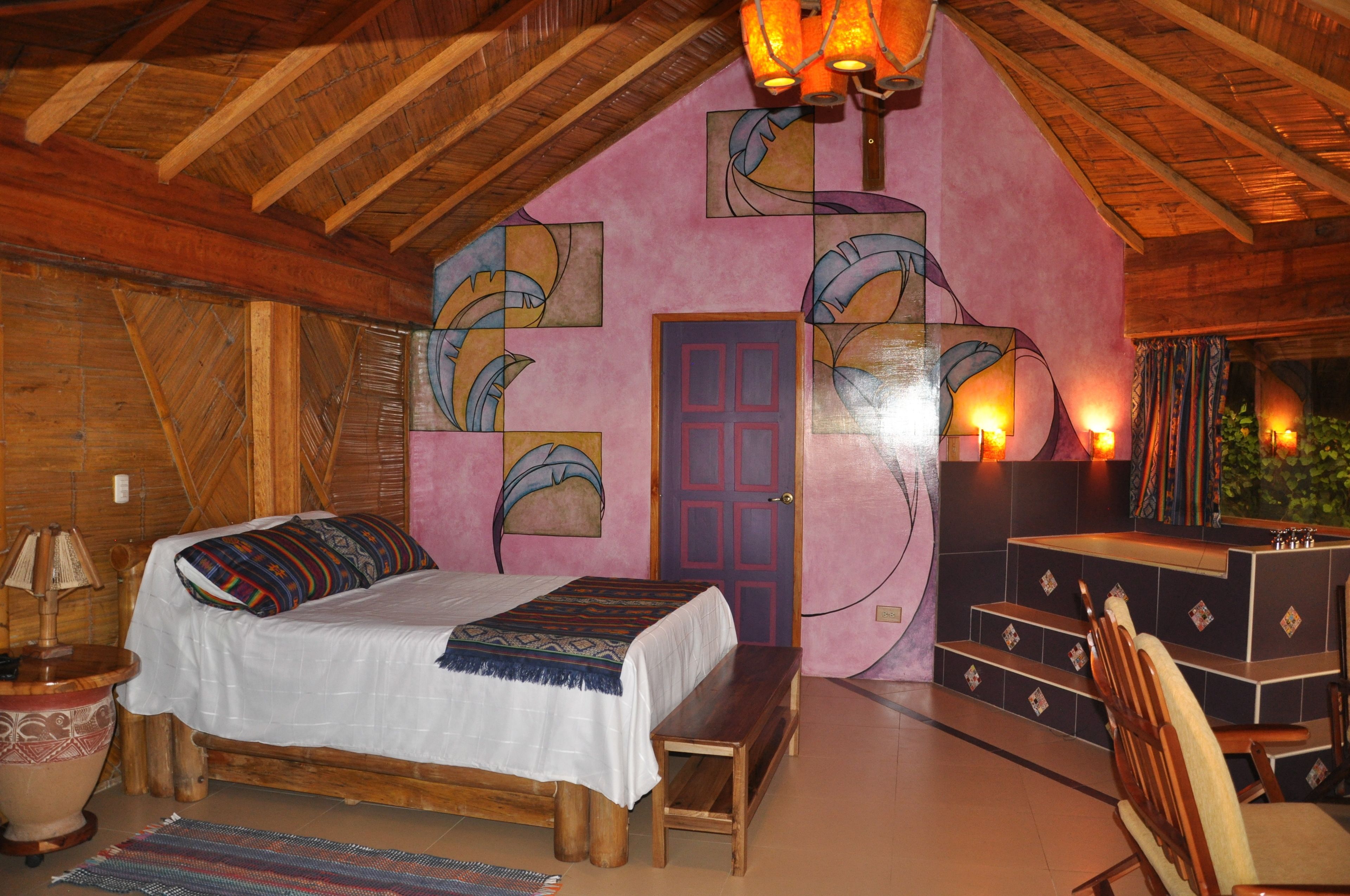 Quarto aconchegante com teto de madeira, cama com manta colorida, um banquinho, escrivaninha com cadeira e porta em parede rosa com obras de arte.