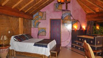 Quarto aconchegante com teto de madeira, cama com manta colorida, um banquinho, escrivaninha com cadeira e porta em parede rosa com obras de arte.