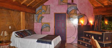 A cozy bedroom with a wooden ceiling, a bed with a colorful blanket, a bench, a desk with a chair, and a door with a pink wall and artwork.