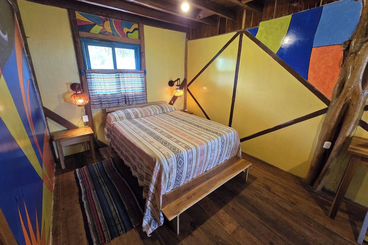 A room with a bed, a window with curtains, a wall with geometric patterns, and a wooden ceiling.