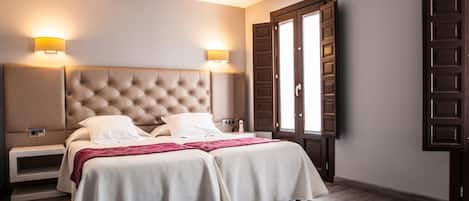 Un dormitorio con cabecera capitonada, dos mesitas de noche, cama con ropa blanca y un amplio ventanal con contraventanas de madera.