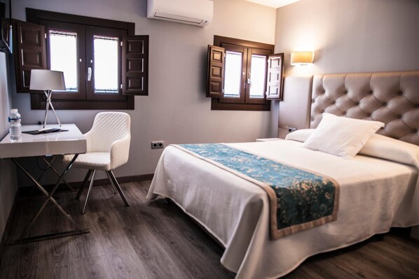 Habitación de hotel con cama, escritorio, silla y dos ventanas con contraventanas.