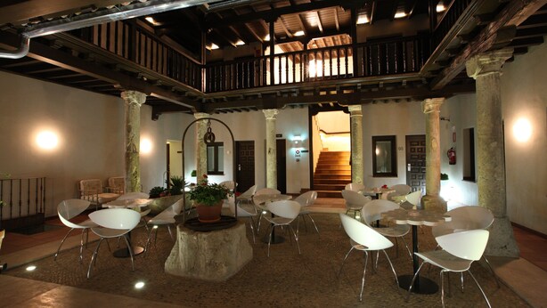 A spacious indoor area with a high ceiling, wooden beams, and a central staircase leading to an upper level. There are several white chairs and tables arranged around a central stone feature with a potted plant. - Evenia Alcalá Boutique Hotel (Alcala de Henares)