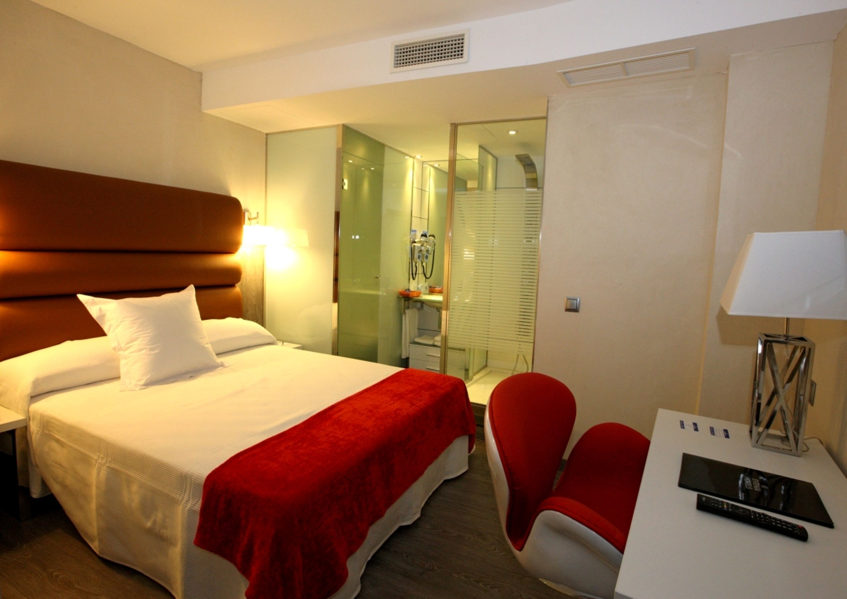 A hotel room with a bed, a desk with a remote control, a chair, and a mirror.
