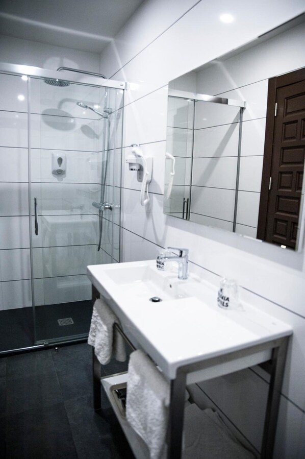 A modern bathroom with a glass-enclosed shower, a white sink with a mirror above it, and a towel rack. - Evenia Alcalá Boutique Hotel (Alcala de Henares)