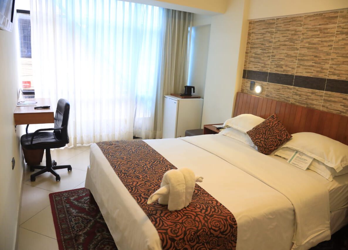 Una habitación de hotel con cama, escritorio, silla, un pequeño armario y ventana con cortinas.