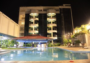 A swimming pool with a covered area, surrounded by greenery and a modern building with multiple balconies.