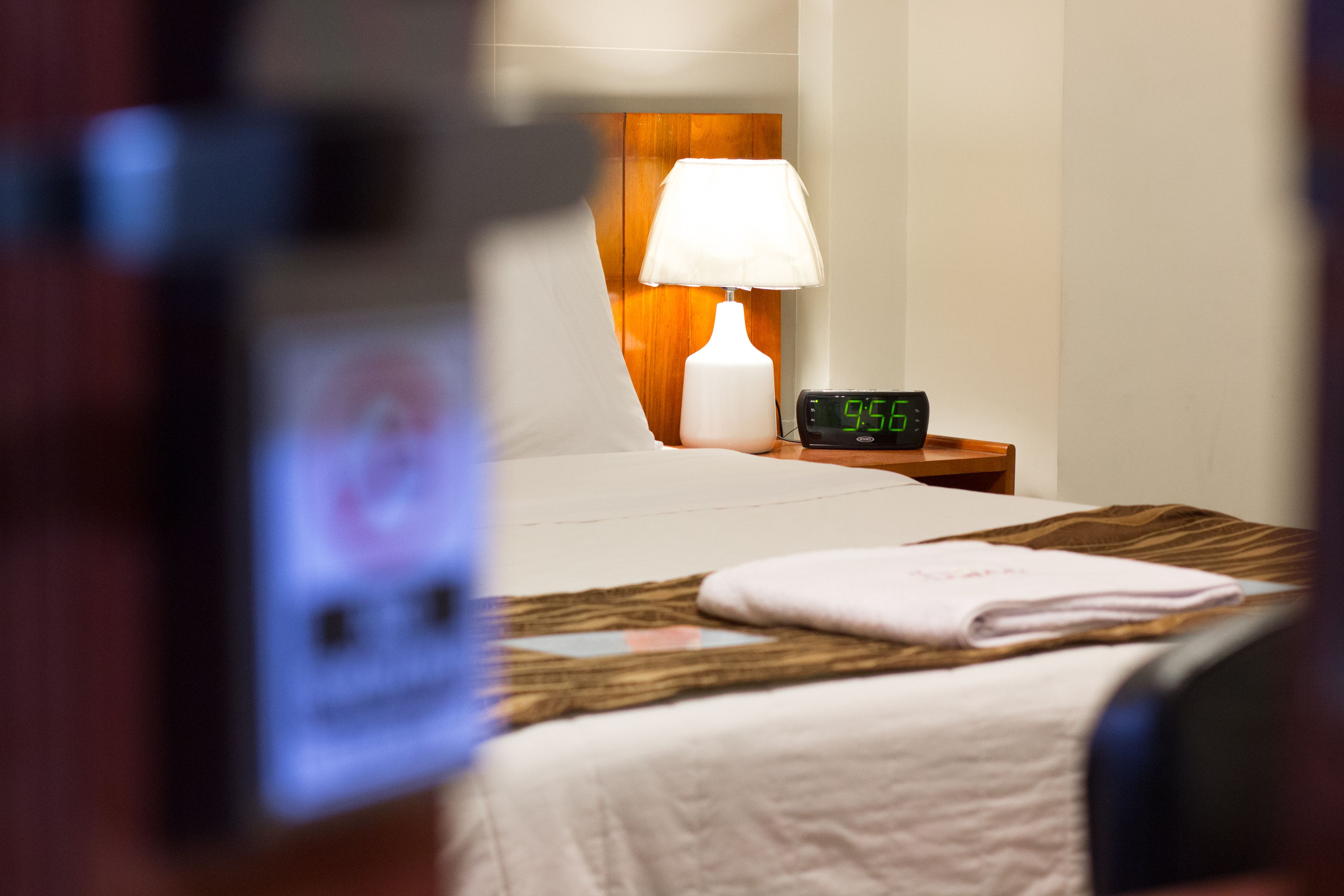 Hotellihuone, jossa on sänky, yöpöytä lampun ja digitaalisen kellon kanssa sekä taitettu pyyhe sängyllä.