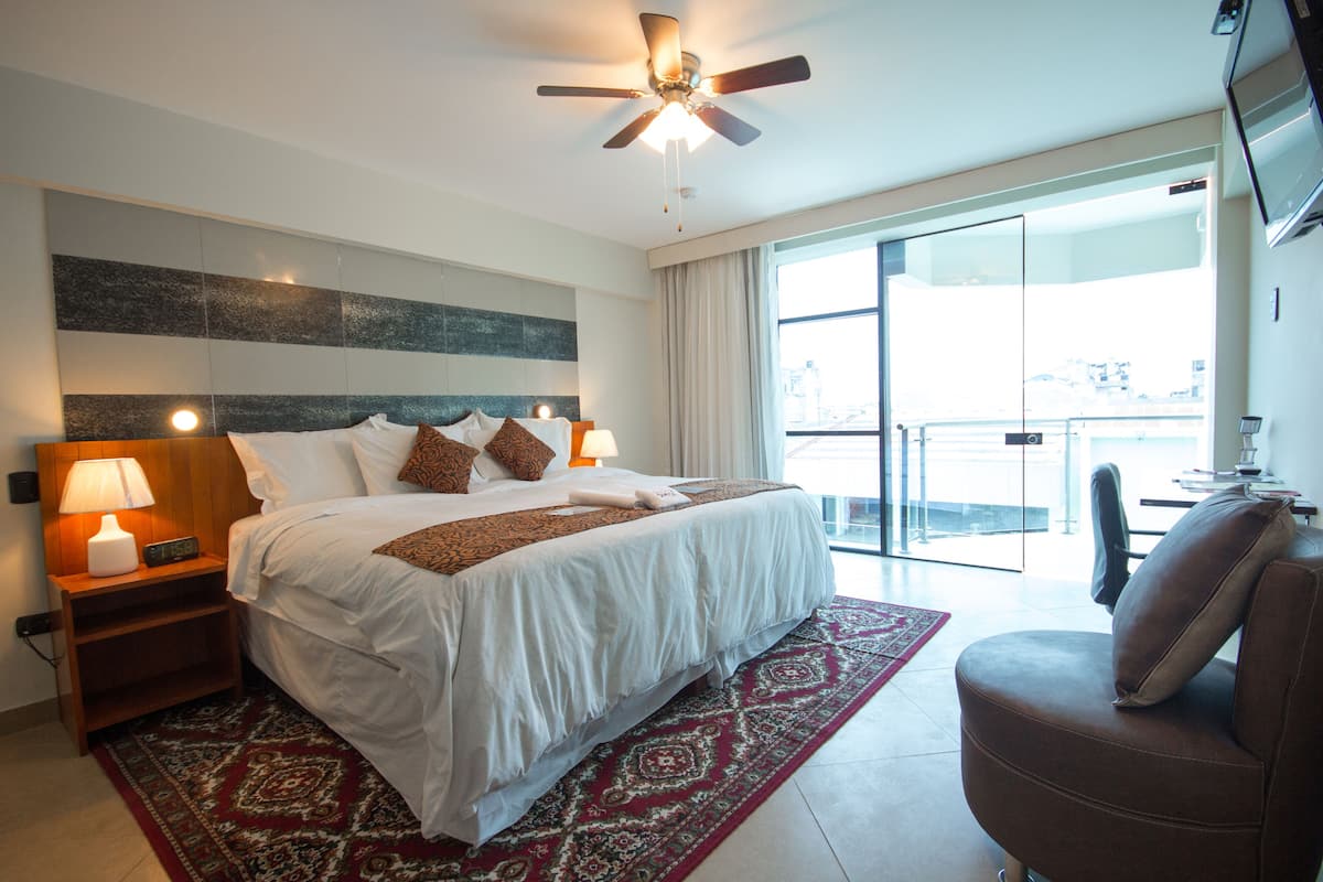 A hotel room with a large bed, a ceiling fan, a TV, and a view of the city through the window.