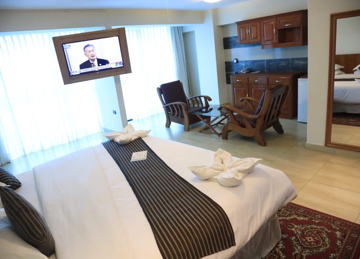 Habitación de hotel con cama, televisor en la pared, zona de comedor con sillas y kitchenette.