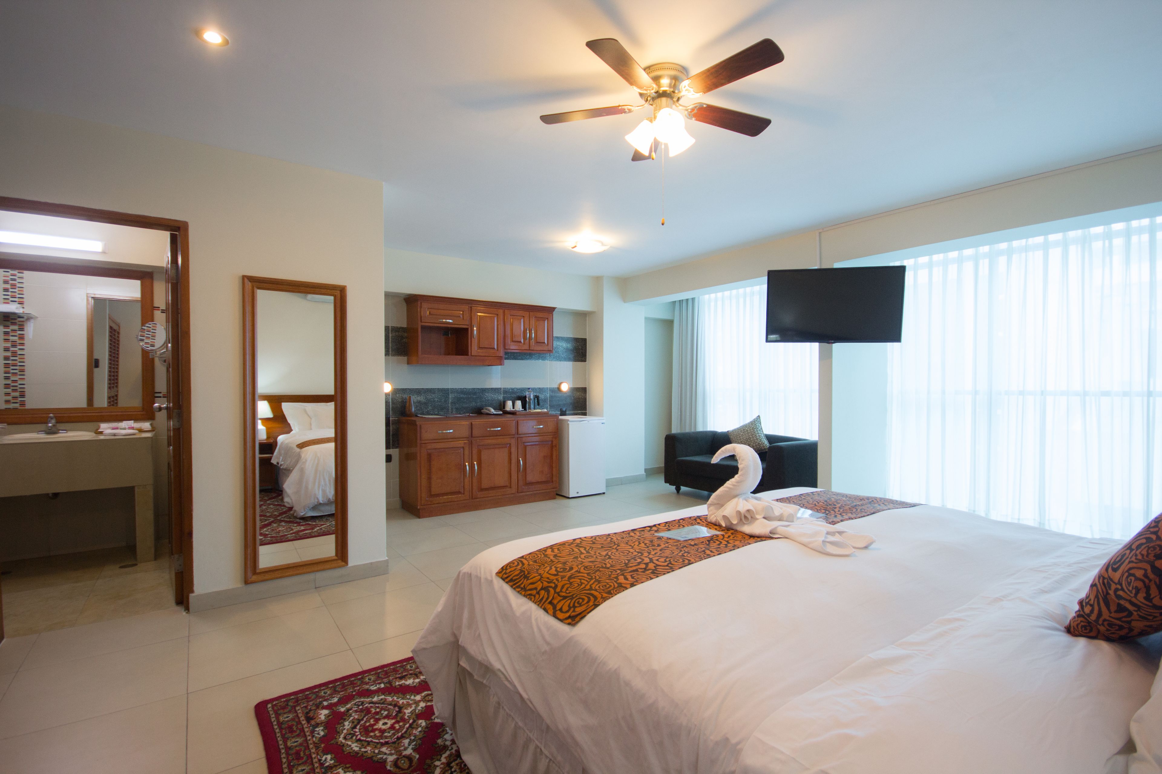Habitación de hotel con cama, ventilador de techo, televisión, baño con espejo y ventana con persianas.