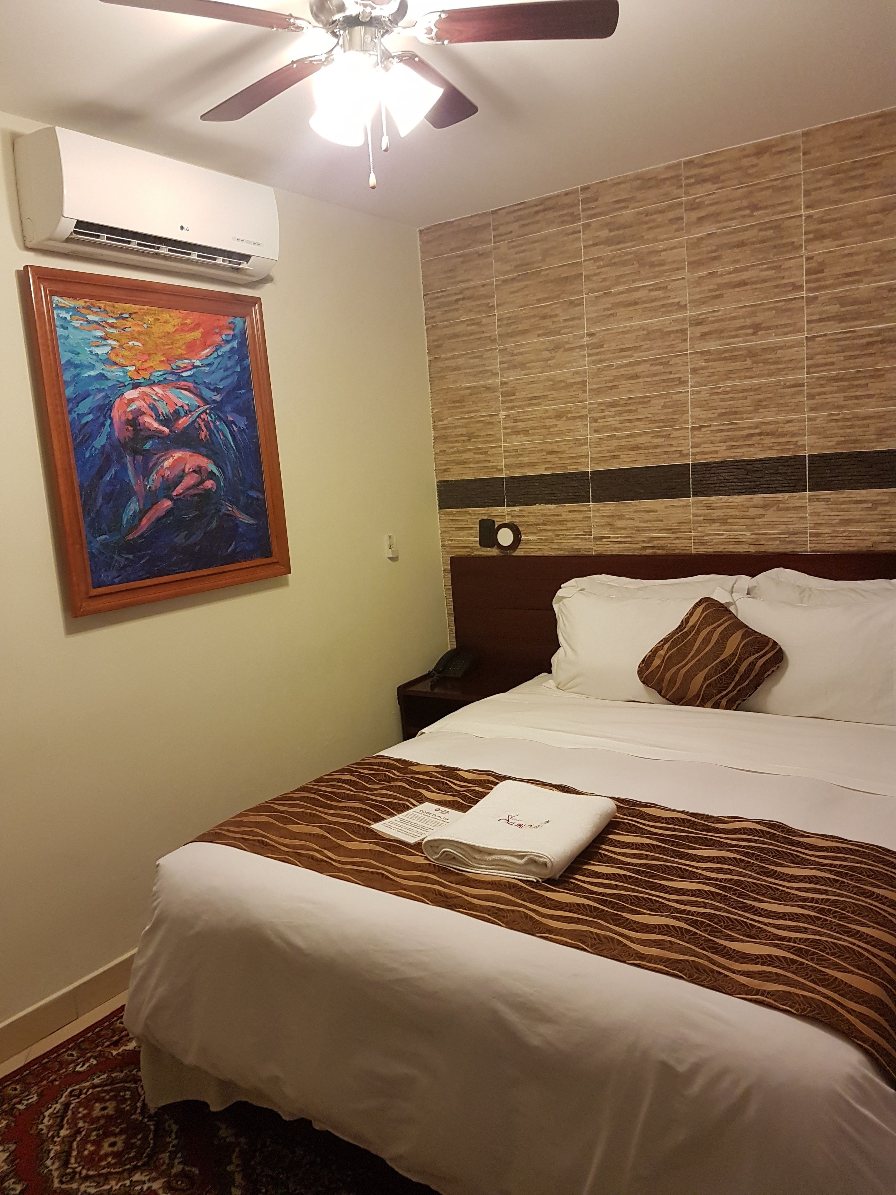Une chambre d’hôtel avec un lit, un ventilateur de plafond, un climatiseur, un tableau, une tête de lit en bois et un tapis à motifs.