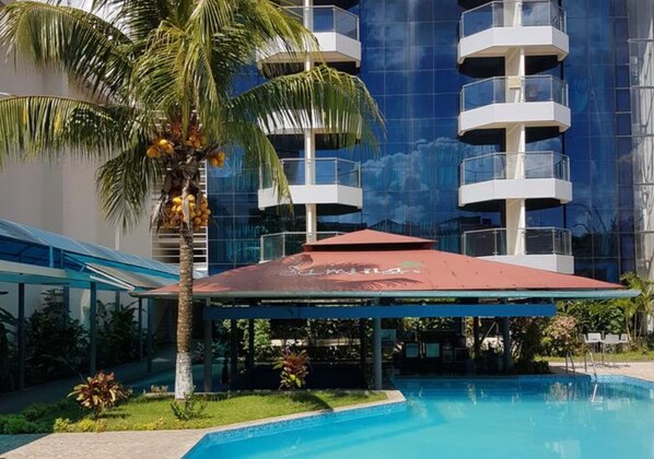 A modern hotel with a swimming pool, palm trees, and a covered area with a red roof.