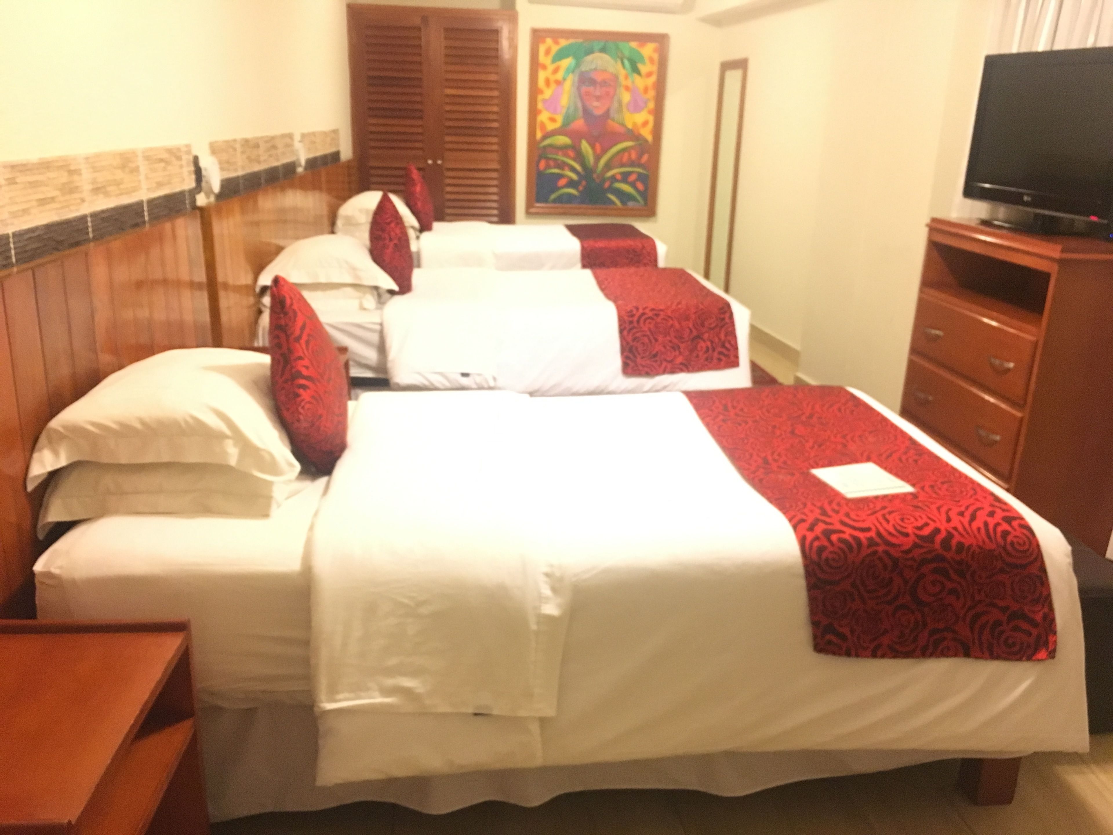 A hotel room with three beds, wooden headboards, a television on a wooden stand, and a colorful painting on the wall.
