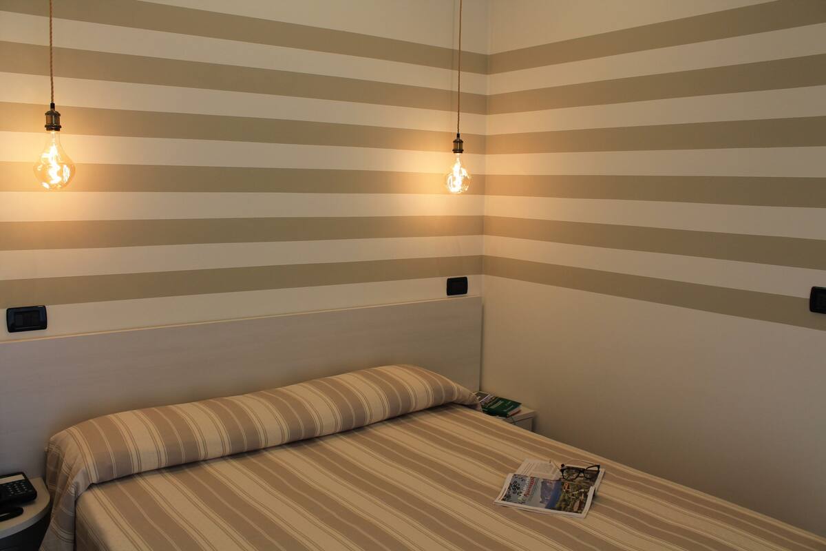 A hotel room with striped walls, a single bed with a striped pillow, two bedside tables, and two hanging light bulbs.