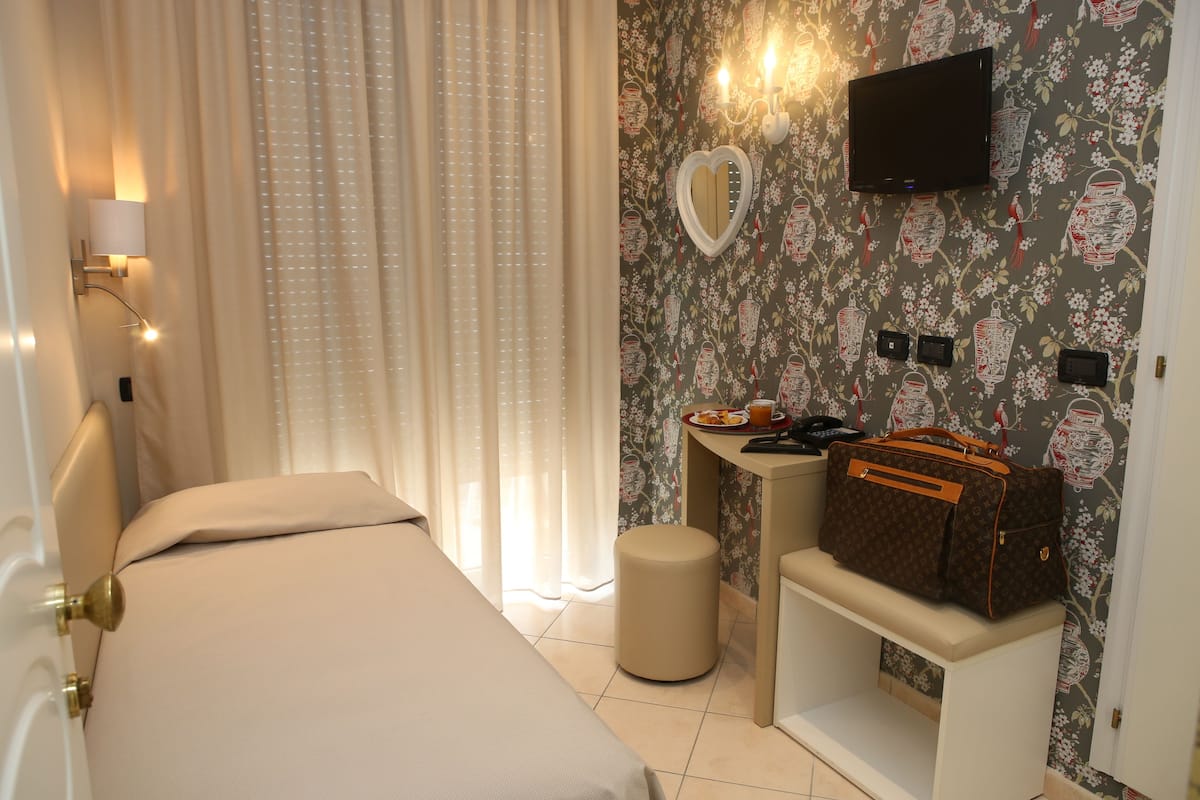 A hotel room with a bed, a desk with a chair, a small stool, a television mounted on the wall, and a suitcase on the desk.