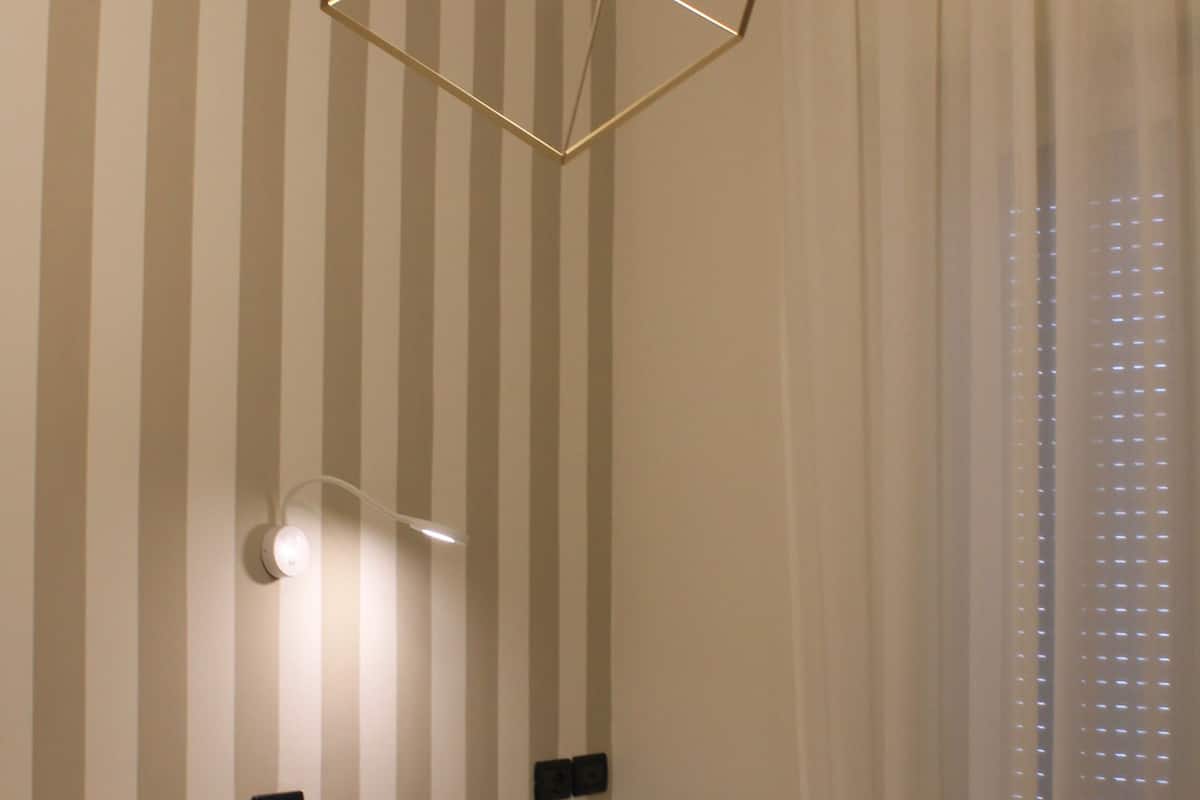 A room with a bed, a geometric light fixture, a wall-mounted switch, and a window with blinds.