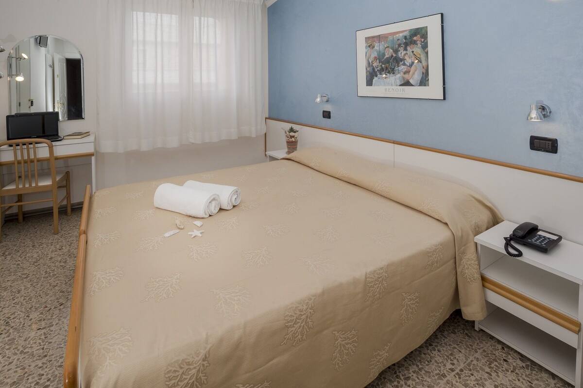 A hotel room with a bed, a desk, a chair, a phone, and a framed picture on the wall.