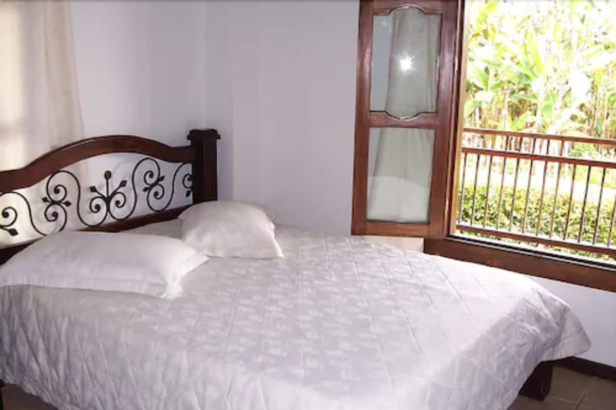 Una cama con cubrecama y almohadas blancas, cabecera de madera con detalles decorativos y ventana con vistas a zonas verdes.