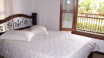 A bed with a white bedspread and pillows, a wooden headboard with decorative elements, and a window with a view of greenery.