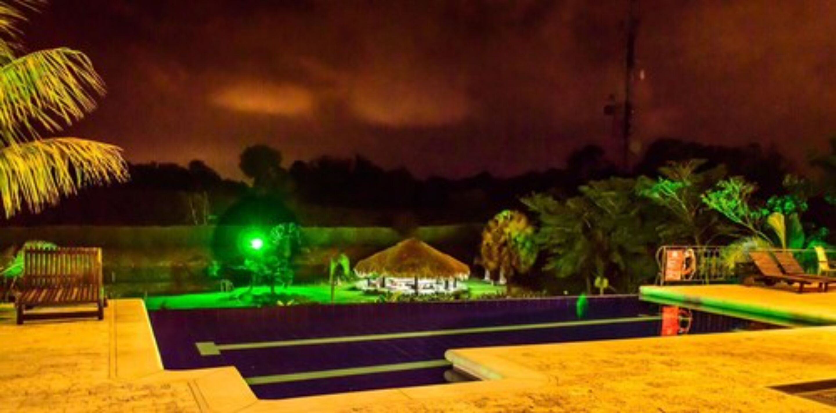 A nighttime view of a resort with a pool, lounge chairs, and a thatched-roof building.