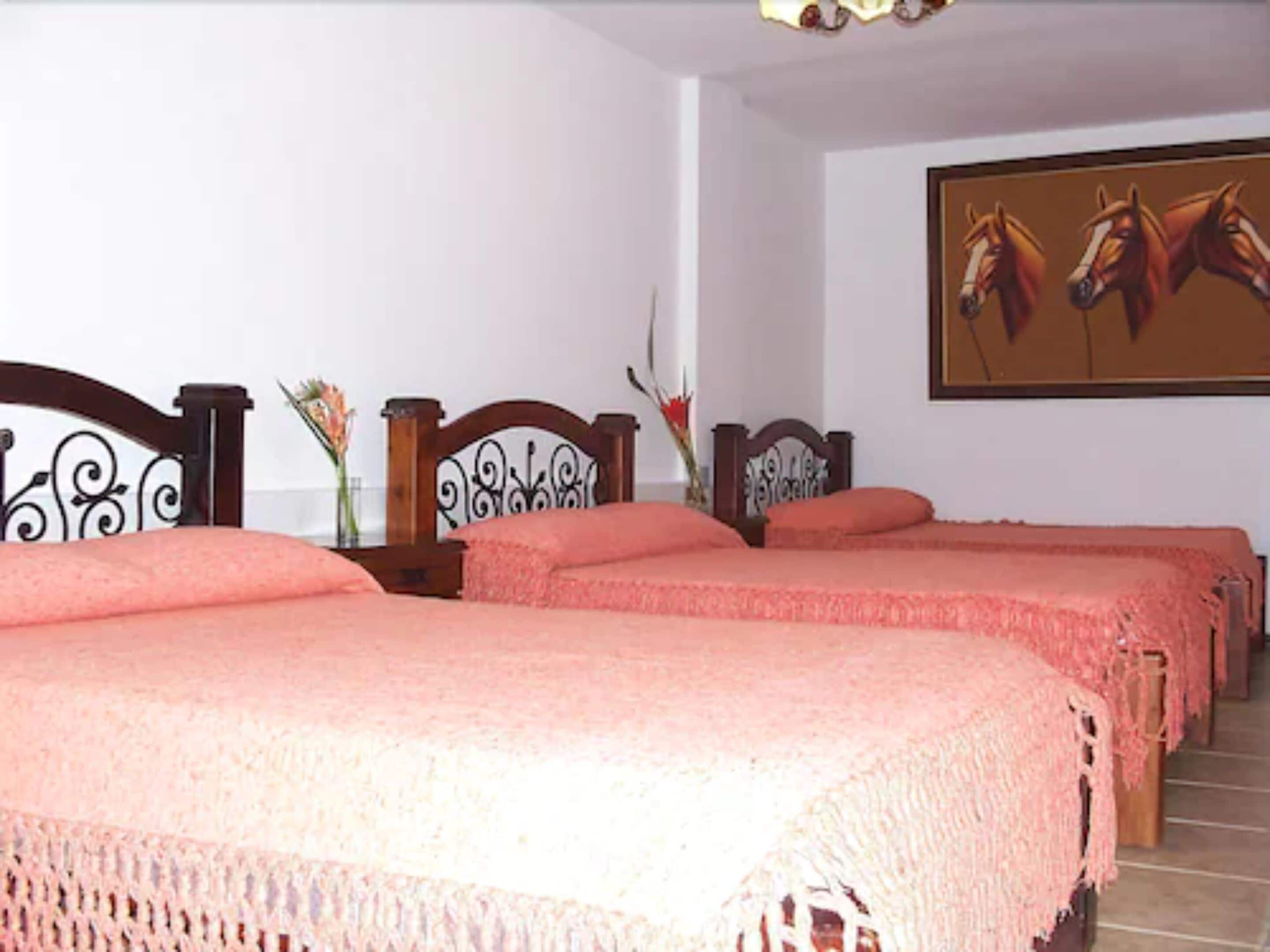 Quarto de hotel com quatro camas, cada uma com um cobertor rosa. Há vasos decorativos nas mesas de cabeceira e um quadro com cavalos na parede.