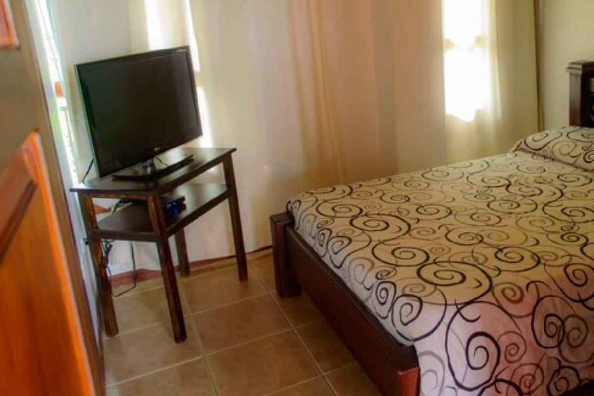 Un dormitorio con una cama, un televisor sobre un soporte de madera y cortinas.