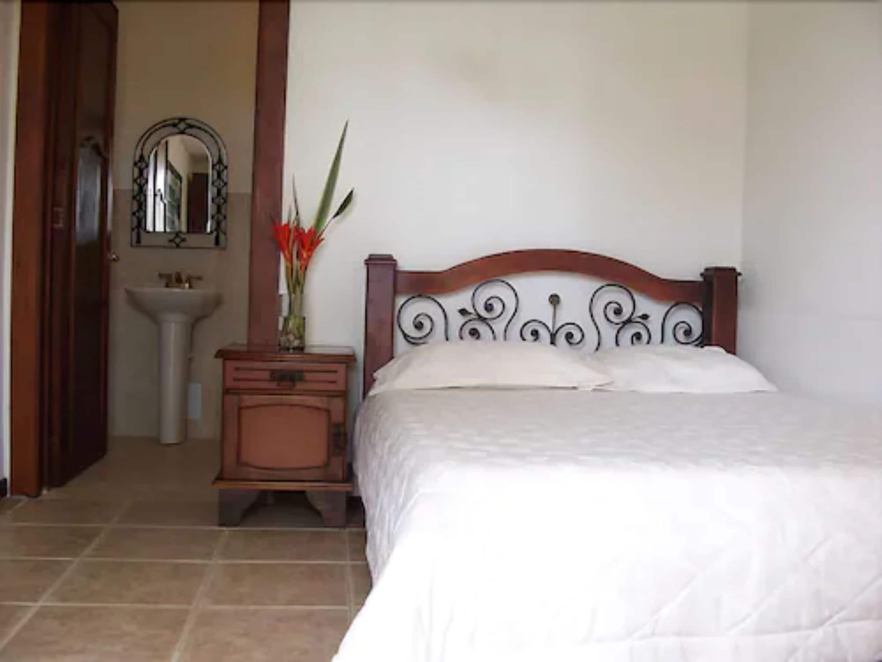A bedroom with a wooden headboard, a bedside table with a vase of flowers, a white bedspread, and a bathroom with a sink and mirror.